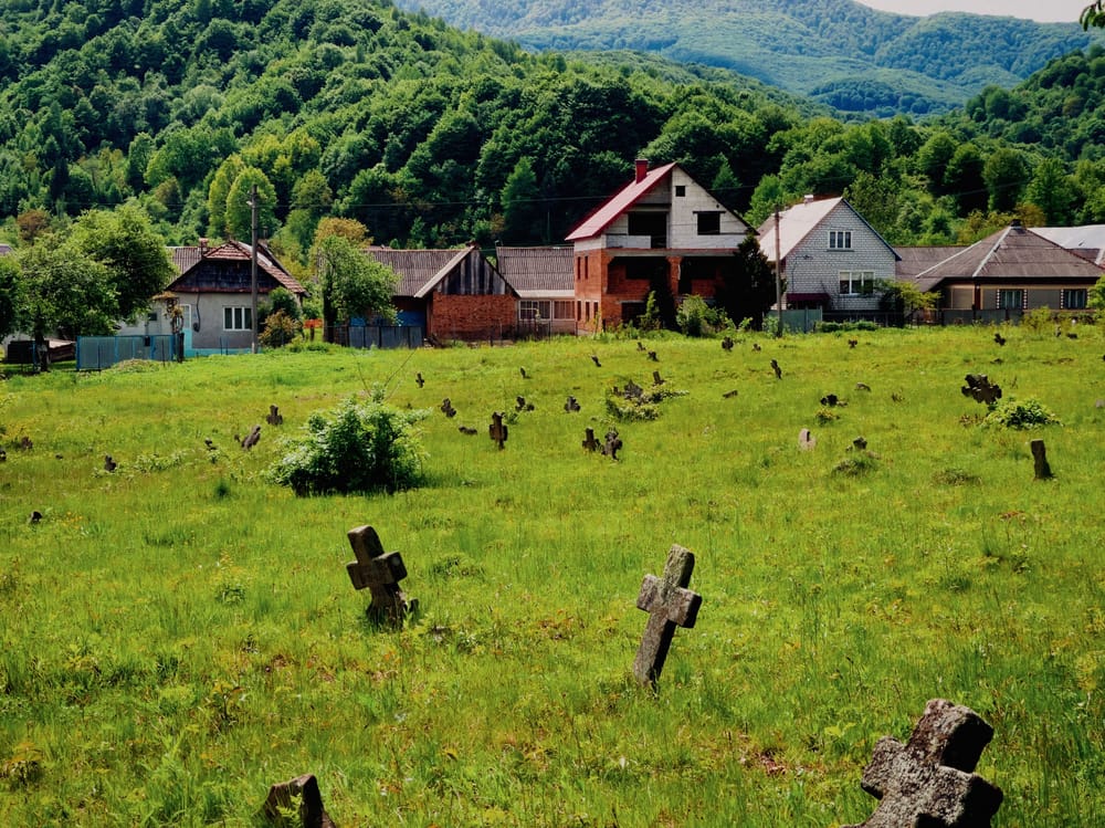 У зеленому польови старі камінні хресты, уд плыну часу вже похылені, а подаґде и втоплені у землю; на позадині людські хыжі, а за нима — складені в дакулько слоюв горы